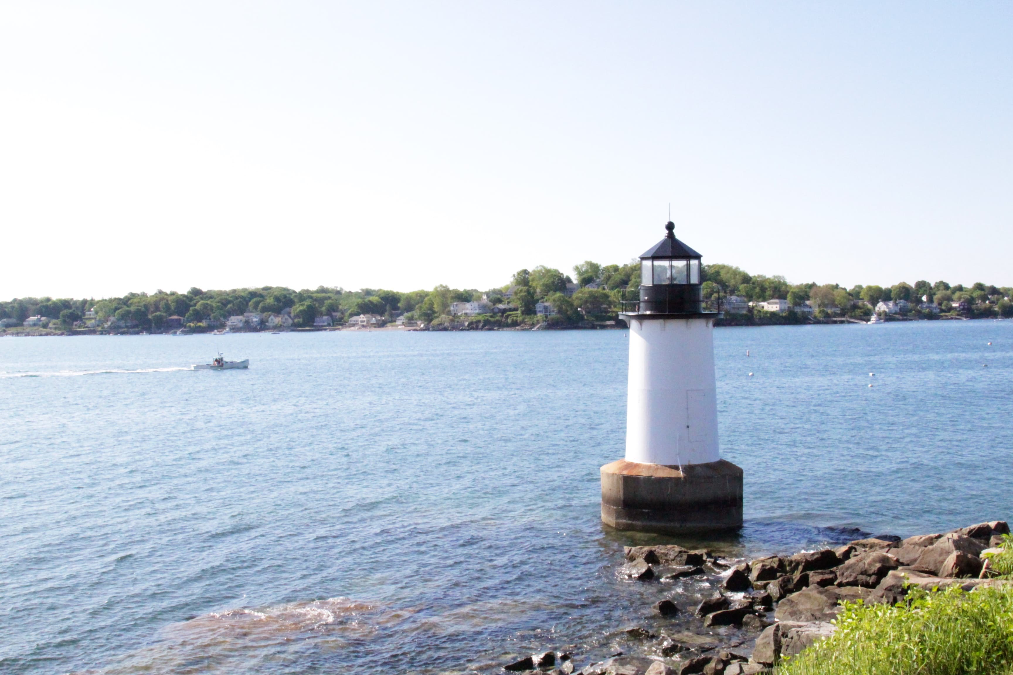 White Fort Pickering lighthouse beside rocks and Salem Harbor, with a boat and wooded shore beyond