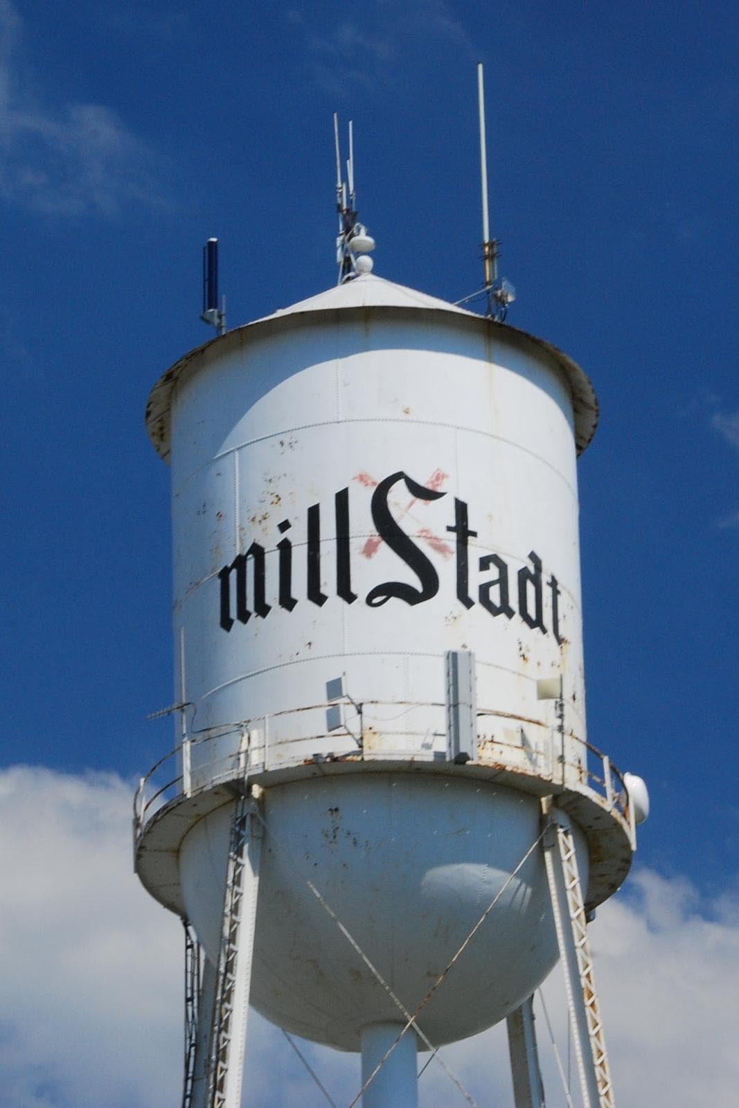 Torre de agua blanca de Millstadt con el nombre de la localidad y antenas bajo cielo azul